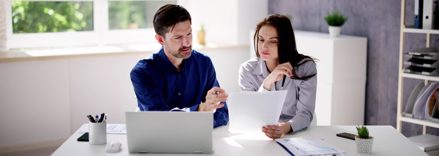 A couple wearing professional business attire sits at a white desk with a computer while reviewing paperwork.